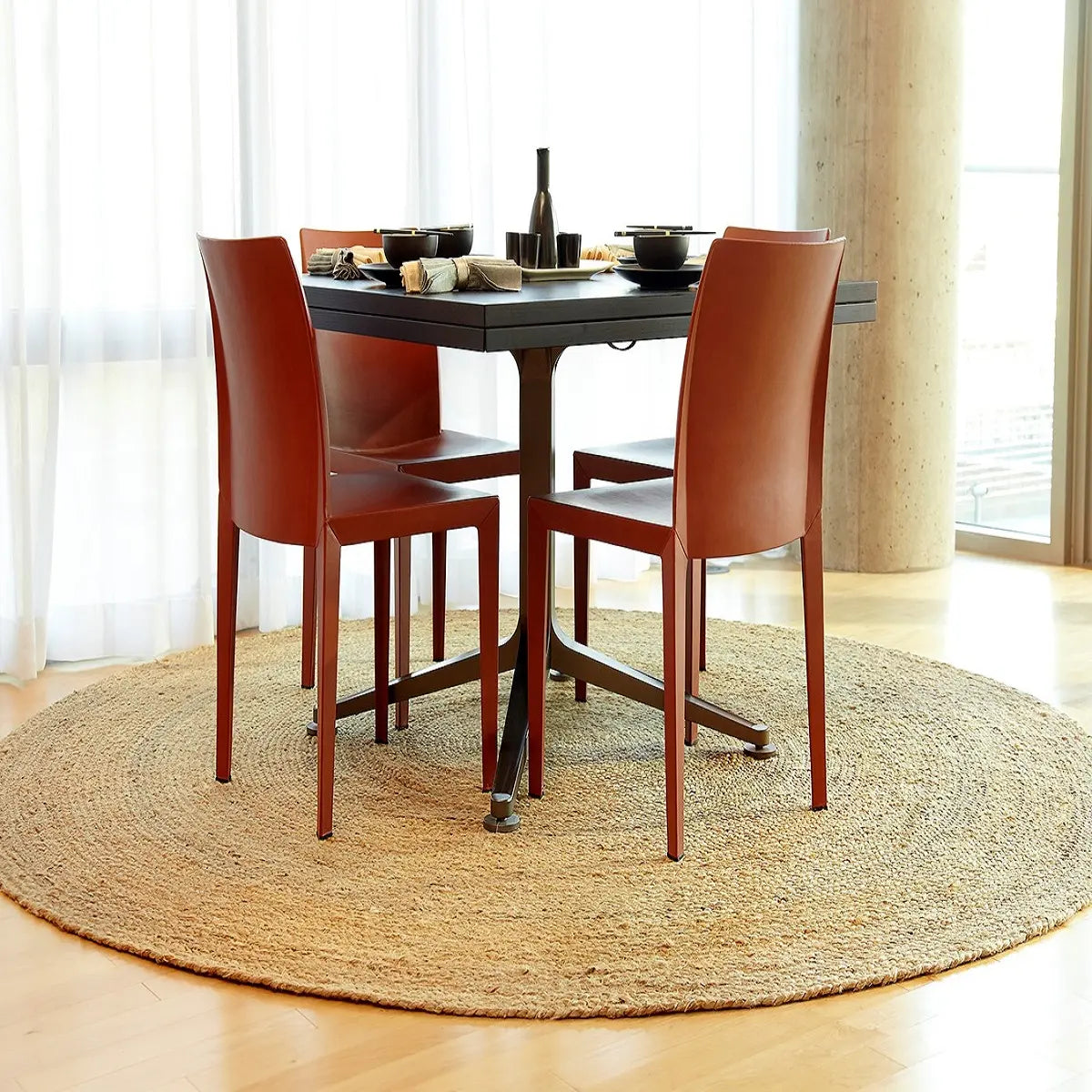 Dining table with red chairs on a round jute rug in a bright room.