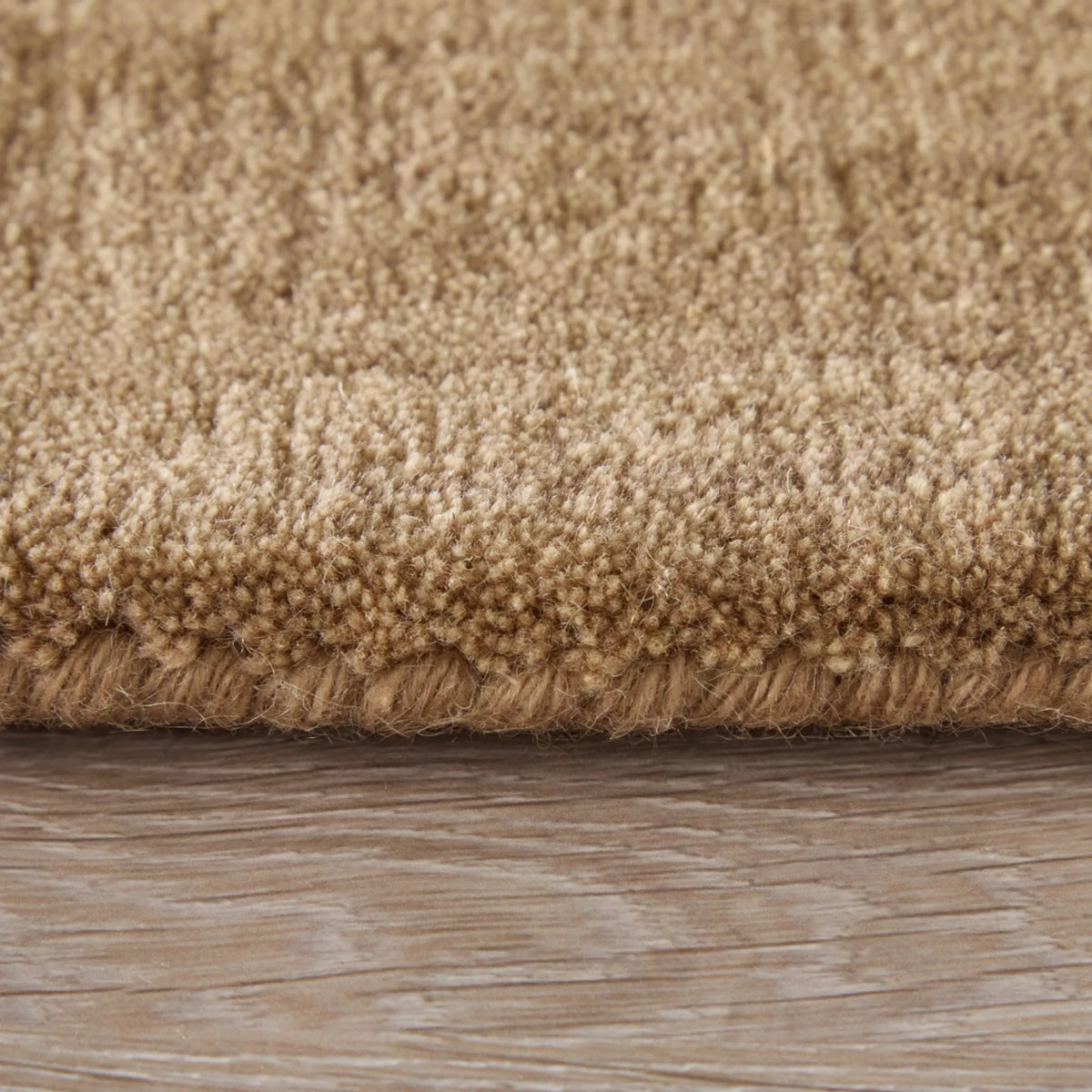 Close-up of a beige shag rug with a thick looped pile and fringed edge, shown resting on a light wood floor.