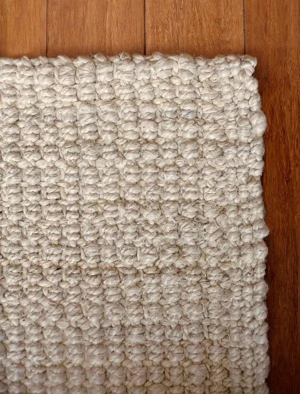 Close-up of a textured ivory rug corner on a wooden floor.