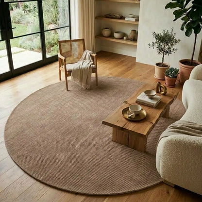 A large round natural fiber area rug in a warm taupe-brown color, styled in a bright, modern living room with a wooden coffee table, a woven armchair, and potted plants.